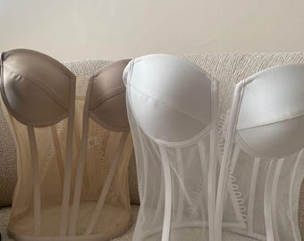 May include: Two corsets, one in a light beige and the other in white, are displayed. The beige corset has a satin cup and a sheer mesh body. The white corset has a satin cup and a sheer mesh body. Both corsets have vertical boning.