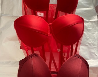 May include: A red corset with three layers of cups. The cups are made of a smooth, satin-like fabric and are lined with a sheer mesh fabric. The corset has a boned structure and is designed to cinch the waist.
