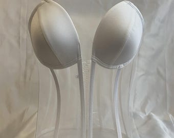 May include: A white corset with a clear, structured body and white trim. The top features two padded cups. The corset has a boned structure and a lace-up back. The design is a combination of opaque and transparent materials.