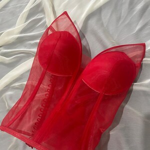 May include: Red mesh corset with a busk closure and boning. The corset has a sweetheart neckline and is made of a sheer fabric.