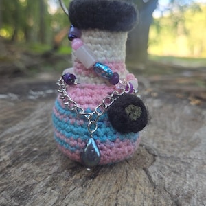 May include: A crochet keychain shaped like a potion bottle with a dark brown felted top. The body features horizontal stripes of white, pink, and blue yarn, adorned with a silver chain, assorted purple and pink beads, and a dark teardrop charm.