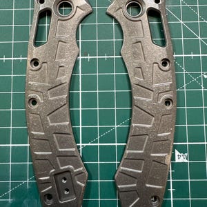 May include: Two gray metal knife handle scales with a textured pattern and multiple holes for screws. The scales have a curved shape and are designed to fit a folding knife. The background is a green cutting mat.