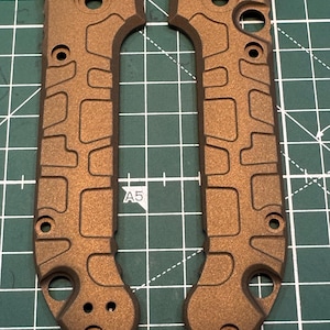 May include: Two bronze-coloured knife handle scales. Each scale has a series of cut-out shapes and holes. The scales are on a green cutting mat with grid lines.