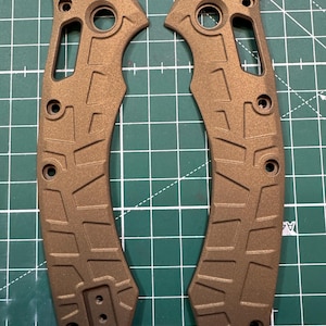 May include: Two bronze-colored knife handle scales with a textured geometric pattern. Each scale has multiple holes for screws and a curved shape. The scales are made of metal and are designed for a folding knife.