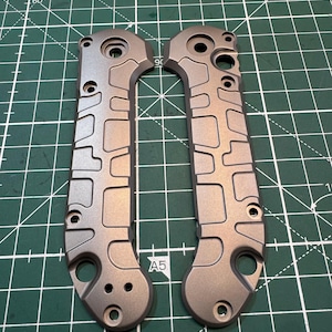 May include: Two grey metal knife handles with a textured, geometric design. Each handle has multiple holes and a curved shape, with a hole at the base. The handles are positioned vertically on a green cutting mat.