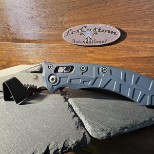May include: A gray and silver folding knife with a serrated edge and a textured handle. The knife is resting on a black stand and a gray stone surface. The text "Lex Custom International" is visible.