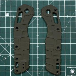 May include: Two dark green metal knife scales with a textured surface and multiple holes. The scales are designed for a folding knife.