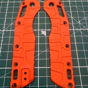 May include: Two bright orange knife handle scales. Each scale has multiple holes and a textured surface. The scales are made of a durable material and are designed to fit a folding knife. The scales are shown on a green cutting mat.