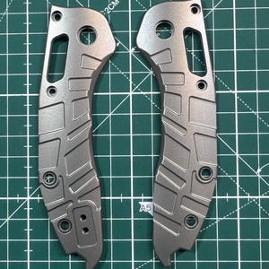 May include: Two gray metal knife scales with a textured pattern. The scales have a hole in the top and a cutout on the side. The scales are on a green cutting mat.