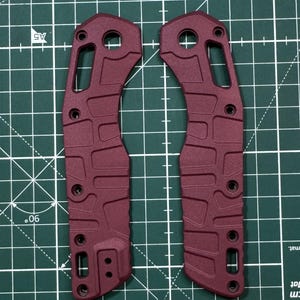 May include: Two burgundy-colored metal knife scales with a textured surface and multiple holes. The scales are designed for a folding knife.