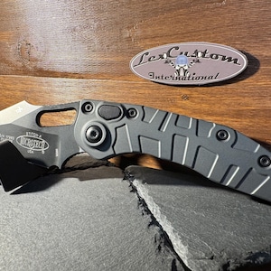 Könnte beinhalten: Ein graues Microtech Stitch-A Messer mit einem schwarzen Taschenclip. Die Klinge ist teilweise geöffnet und zeigt die scharfe Schneide. Der Griff hat ein strukturiertes Design. Das Messer liegt auf einer dunkelgrauen Oberfläche, mit einem LexCustom International Logo im Hintergrund.