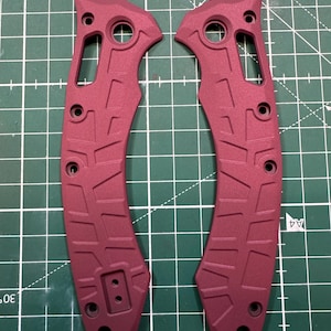 May include: Two burgundy-colored knife handle scales. Each scale has a contoured shape with multiple holes and a textured pattern. The scales are made of a durable material and are designed for a secure grip. The scales are shown on a green cutting mat.