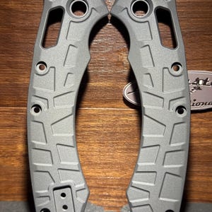 May include: Two gray knife handle scales with a textured geometric pattern. Each scale has multiple holes and a cutout section. The scales are made of a durable material and are designed for a secure grip.