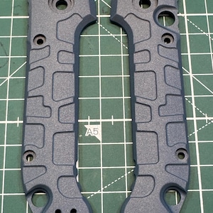 May include: Two grey knife handle scales with a textured, geometric pattern. Each scale has multiple holes for screws and a curved shape for a comfortable grip. The scales are displayed on a green grid.