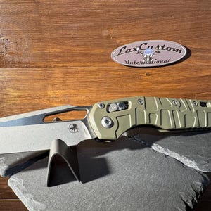 May include: A folding knife with a silver blade and an olive green handle. The knife is displayed on a dark gray stone surface. The handle has a textured design. A logo that says "Lex Custom International" is in the background.