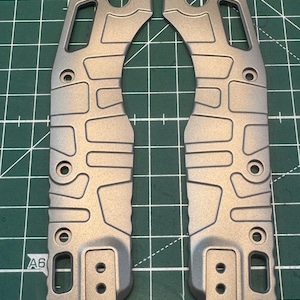 May include: Two gray metal knife scales with a series of holes and cutouts. The scales have a textured surface and a curved design. The parts are designed to be assembled together to form a knife handle.