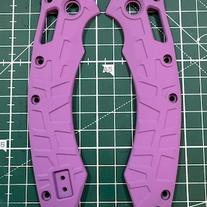 May include: Two lavender-colored knife handle scales. Each scale has a curved shape with multiple holes and a textured surface. The scales are designed to fit together, forming a complete handle. The background is a green cutting mat.