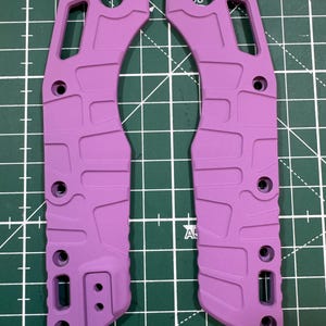May include: Two matching, lavender-colored knife handle scales. Each scale has a curved shape with multiple holes and a textured surface. The scales are designed to fit together, forming a complete handle.