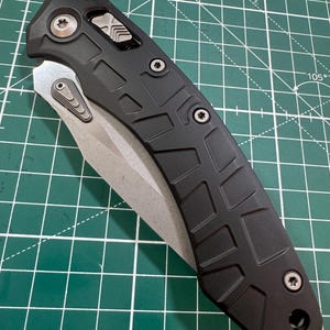 May include: A black folding knife with a silver blade. The handle has a textured, geometric pattern and is secured with silver screws. The blade is partially open, revealing its sharp edge. The knife is on a green cutting mat.