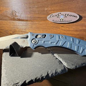 May include: A blue and silver folding knife with a textured handle and a partially serrated blade. The knife rests on a dark gray stone surface. The blade is approximately 3.5 inches long. The knife has the text "LexCustom International" on the wooden background.