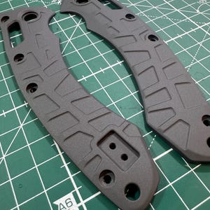 May include: Two gray knife handle scales with a textured geometric pattern and multiple holes for screws. The scales are lying on a green cutting mat with white grid lines.