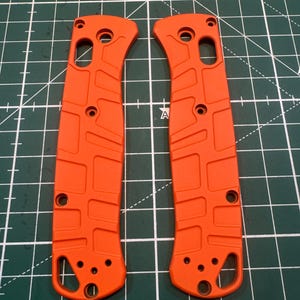 May include: Two bright orange knife handle scales. Each scale has a series of holes and a textured, rectangular pattern. The scales are curved and designed to fit the shape of a knife handle. The scales are shown against a green cutting mat.