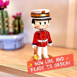May include: A small figurine of a person in a red military-style uniform with white trousers, a white cap with gold trim, and a gold instrument in one hand. A red rectangular sign in the foreground features the text: NOW LIVE AND READY TO ORDER!