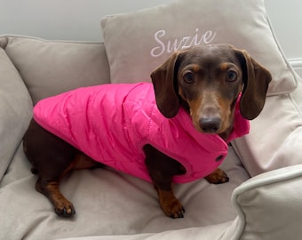 May include: A short-haired brown and black dachshund wearing a bright neon pink quilted puffer vest. The dog is positioned in a light beige pet bed that features a matching pillow with the name Suzie embroidered in white cursive script.