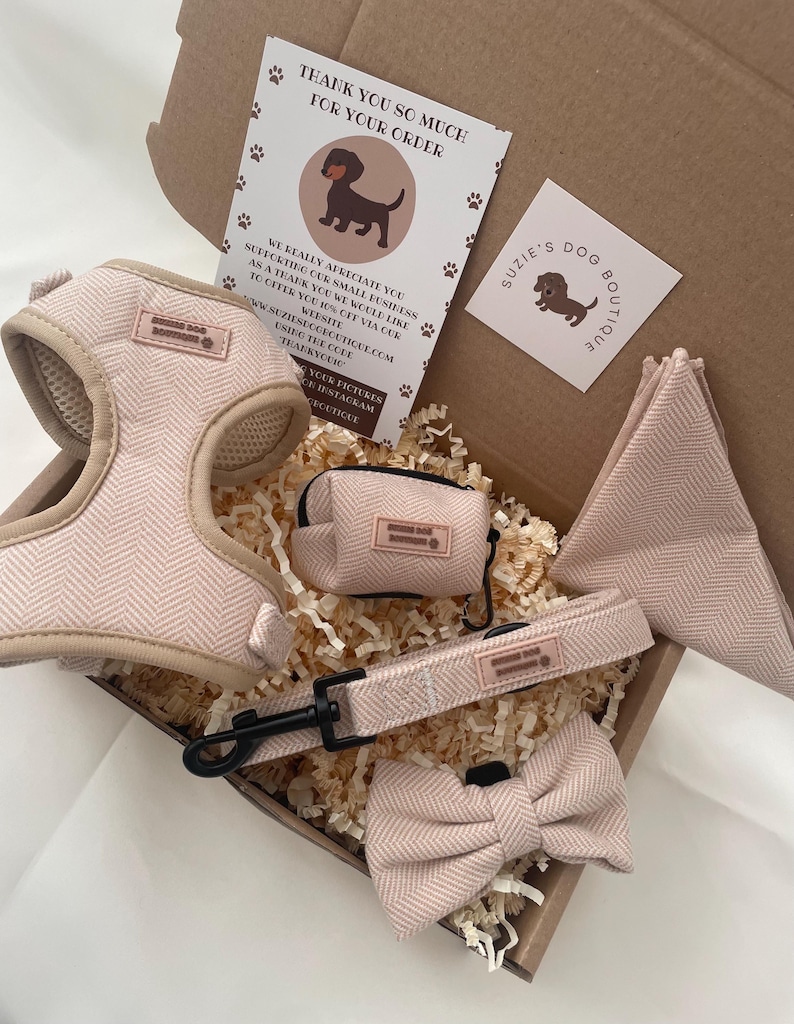 Puede incluir: Un conjunto de accesorios para perro en una caja de cart&oacute;n, que incluye un arn&eacute;s de color rosa claro con patr&oacute;n de espiga, una correa a juego con clip de metal negro, un dispensador de bolsas, una pajarita y un pa&ntilde;uelo triangular con etiquetas.