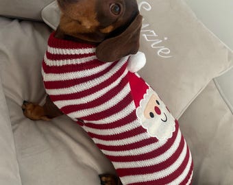 May include: A brown dachshund wearing a red and white striped Christmas sweater with a Santa Claus face on the front. The dog is lying on a light gray cushion, looking upwards. The sweater has a white pom-pom on the back.