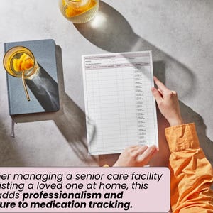 May include: A medication tracking form with a grid layout, held by a person wearing an orange shirt. A blue notebook and a glass of beverage with a gold straw are on the left. The form adds structure to medication tracking.
