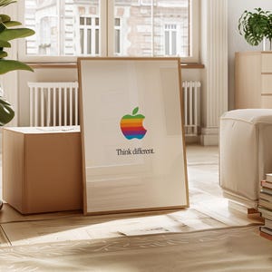 May include: A framed print featuring the Apple logo, a colorful rainbow apple, and the text "Think different." The artwork is displayed in a bright, airy room with a large window and wooden floors. The print is leaning against a cardboard box.