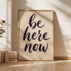 以下が含まれることがあります： 明るいベージュの背景に、濃い紫色の筆文字で「be here now」と中央に書かれた長方形の額入りウォールアート。フレームは明るい色の木材で作られており、シンプルで清潔感のあるデザインが特徴です。