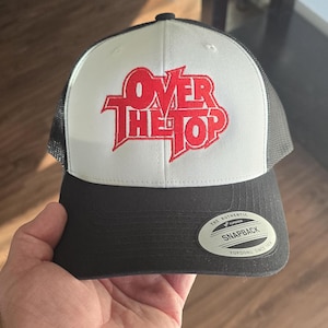 May include: A black and white trucker hat with the text "Over The Top" in red. The hat has a white front panel, a black brim, and a mesh back. The hat has a snapback closure.