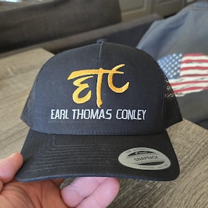 May include: Black trucker hat with a curved brim and mesh back. The hat features the embroidered text "ETC" in gold, above the text "EARL THOMAS CONLEY" in white. The hat has a snapback closure.