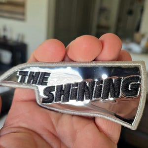 May include: A silver-colored patch with the words "THE SHINING" embroidered in black. The patch has a silver border and a unique shape. The patch is likely a collectible item or a decorative accessory.