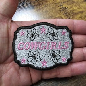 May include: A gray and black embroidered patch with the word "COWGIRLS" in pink lettering. The patch is decorated with pink stars and white flowers.