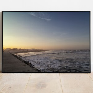 K&ouml;nnte beinhalten: Ein Foto von einem Strand bei Sonnenuntergang mit einem langen, gepflasterten Weg, der zum Wasser f&uuml;hrt. Der Himmel ist ein Farbverlauf von Blau, Orange und Gelb, und die Wellen brechen an der K&uuml;ste.