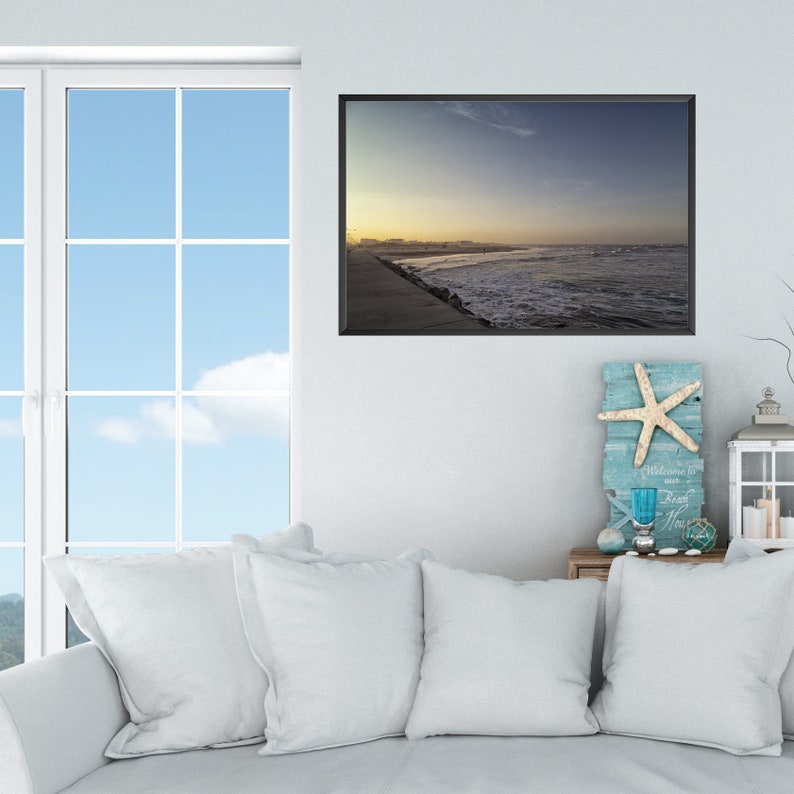 K&ouml;nnte beinhalten: Ein gerahmtes Foto einer Strandszene mit bew&ouml;lktem Himmel und Wellen, die an das Ufer schlagen. Das Foto h&auml;ngt an einer wei&szlig;en Wand &uuml;ber einem wei&szlig;en Sofa mit Kissen.