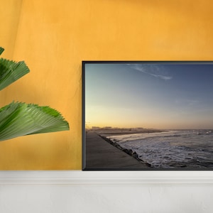 K&ouml;nnte beinhalten: Ein gerahmtes Foto einer Strandszene mit einem langen, gepflasterten Weg, der zum Meer f&uuml;hrt. Der Himmel ist hellblau mit ein paar Wolken, und das Wasser ist tiefblau mit wei&szlig;en Wellen, die an der K&uuml;ste brechen.