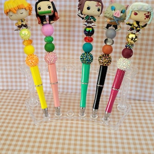 May include: Six colorful pens with anime character toppers and colorful beads. The pens are displayed in a clear plastic holder on a light brown gingham background.