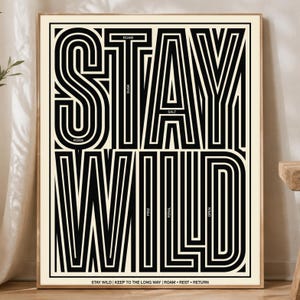 May include: A rectangular wall art print featuring the words STAY WILD in large, bold, black, multi-line typography against a cream background. Smaller text includes the words ROAM, DUSK, SALT, PINE, FERAL, OPEN, and a bottom line of text.