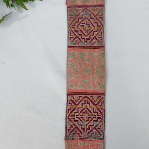 May include: A long, narrow strip of fabric with a repeating pattern of embroidered squares. The squares are in shades of pink, red, green, and blue. The fabric is sewn together with a blue trim at the bottom.