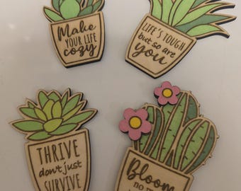 May include: Four wooden succulent and cactus magnets with inspirational quotes. The magnets feature green and light green plants in brown pots. The quotes include "Make your life cozy", "Life's tough but so are you", "Thrive don't just survive", and "Bloom no matter what".