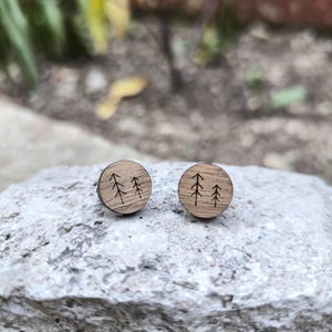 Puede incluir: Dos pendientes de botón redondos de madera. Cada pendiente presenta un diseño tallado de tres árboles estilizados. Los pendientes son de color marrón claro y están sobre un fondo de piedra natural.