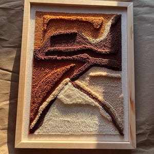 May include: A rectangular punch needle wall art piece framed in light-coloured wood. The textile artwork features abstract, layered shapes in shades of cream, burnt orange, and deep brown, creating a textured, multi-dimensional landscape effect.