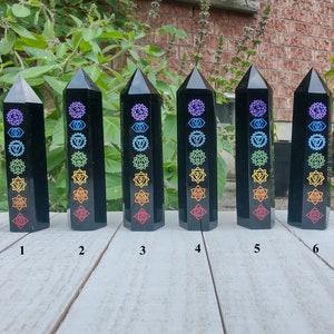 Könnte beinhalten: Sechs schwarze Obsidian-Obelisken mit farbigen Chakra-Symbolen. Jeder Obelisk ist ein hoher, rechteckiger Prismakörper mit spitzer Spitze. Die Chakra-Symbole sind vertikal angeordnet, nummeriert von 1 bis 6.
