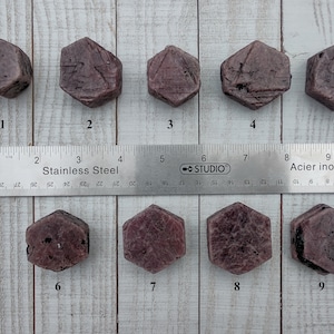 May include: Nine faceted, reddish-purple gemstones are arranged in a row, next to a stainless steel ruler. The ruler measures in inches, with the gemstones ranging from approximately 0.5 to 1 inch in size. The gemstones have a hexagonal shape.