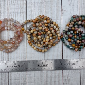 Peut inclure: Trois rangées de perles de pierres précieuses roses, beiges, jaunes, marron, vertes et bleues. Une règle en acier inoxydable avec des mesures en centimètres est placée en dessous, mesurant environ 30 cm.