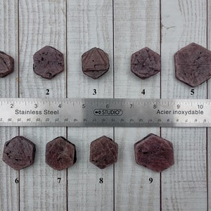 May include: Nine hexagonal, reddish-purple gemstones are arranged in a row. Each stone is unique, with etched geometric designs. A stainless steel ruler with inch markings is placed above the stones, providing scale. The background is a light wood grain.
